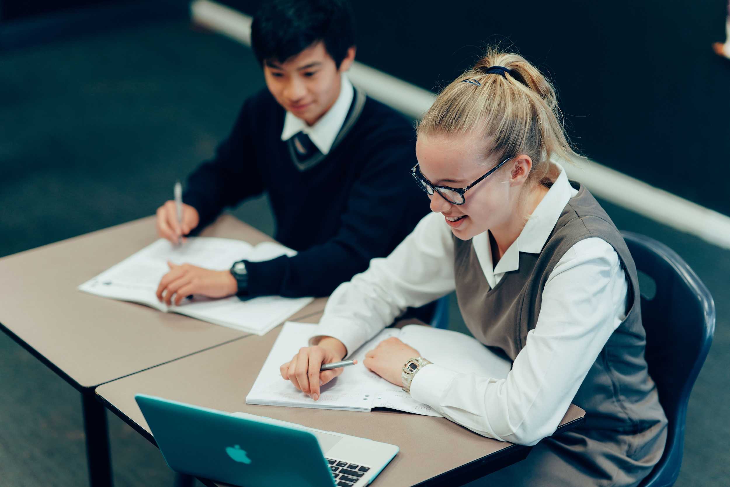 male and female high school student in formal uniform sitting at desk working together on laptop